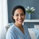 Close-up portrait of a happy woman in her late 20s to early 30s, smiling at the viewer, with a blurred slate blue and white b