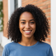 A smiling young woman with dark curly hair looks at the camera. She wears a slate blue top and stands in front of blurred ter